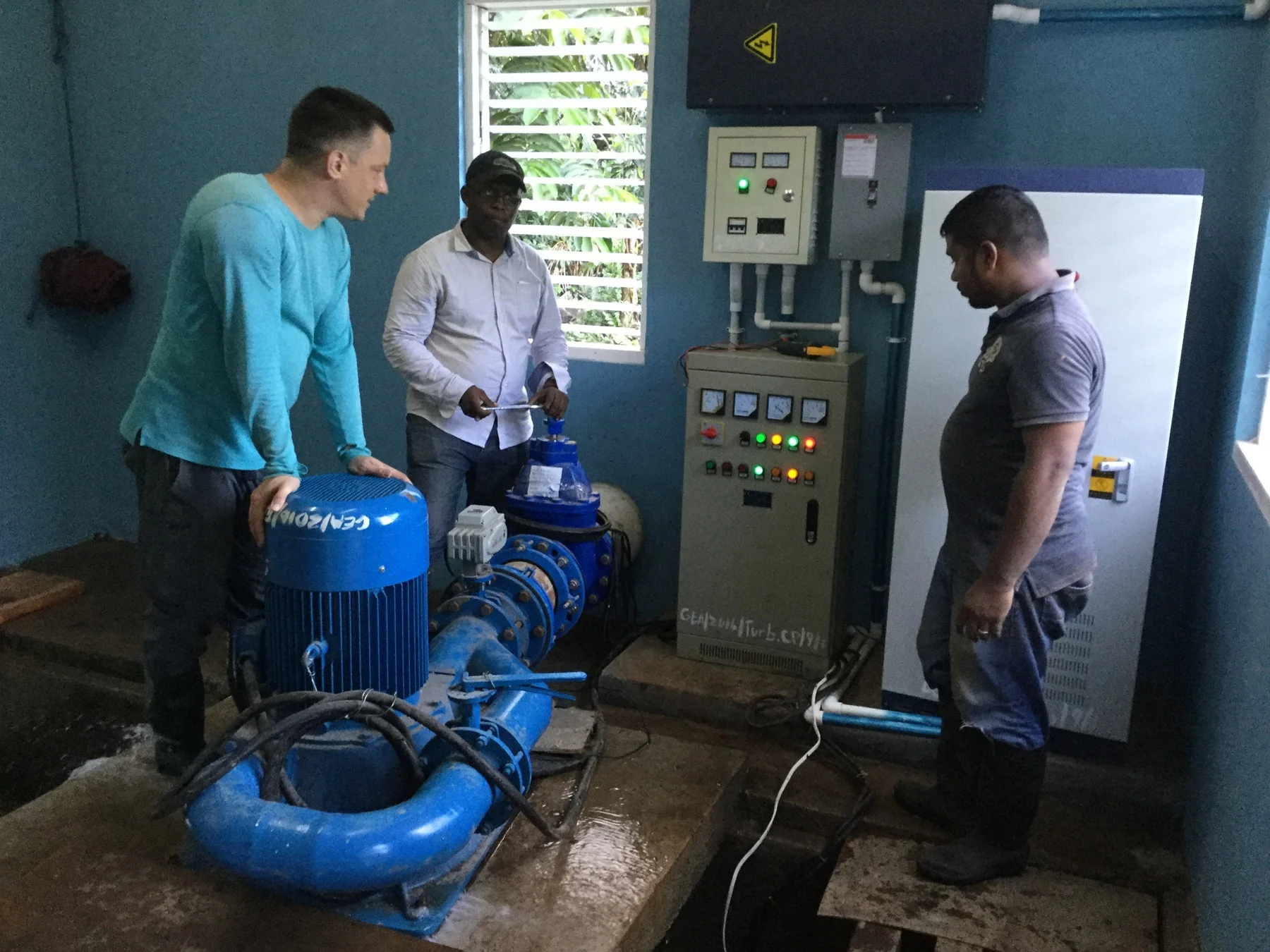Three people beside a small hydropower turbine and control panel inside the Hosororo powerhouse in Guyana